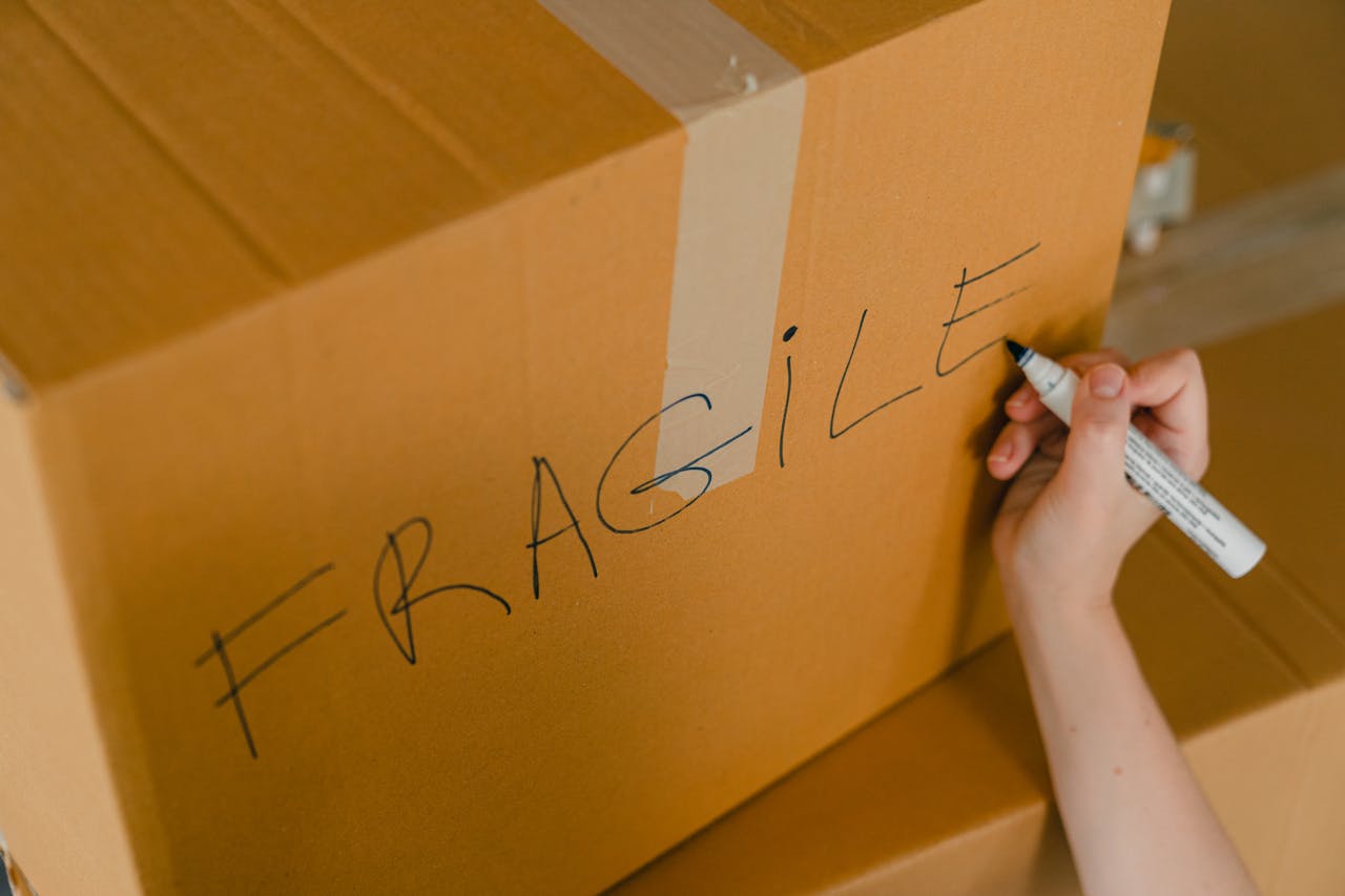 From above of crop unrecognizable person writing with marker on cardboard box with fragile products before relocation